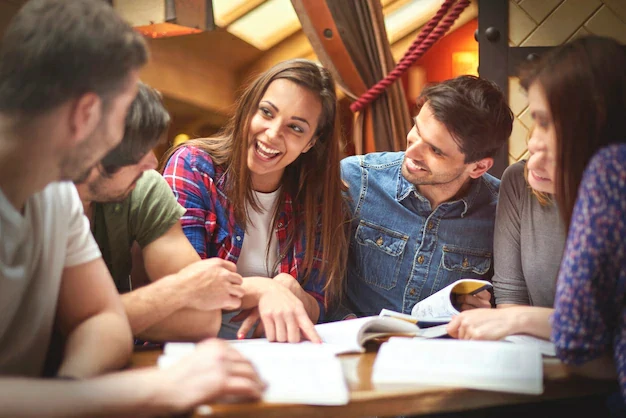 Grupo de estudos em um encontro agradável, com várias pessoas sentadas ao redor de uma mesa com livros, enquanto uma jovem no centro sorri e aponta para algo no livro, simbolizando cooperação e aprendizado coletivo.