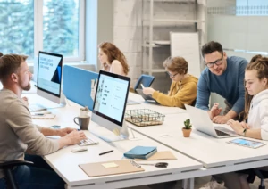 Pessoas trabalhando em computadores dentro de um escritório, com um rapaz auxiliando uma moça, simbolizando ambiente de trabalho tranquilo devido a práticas da NR-1.