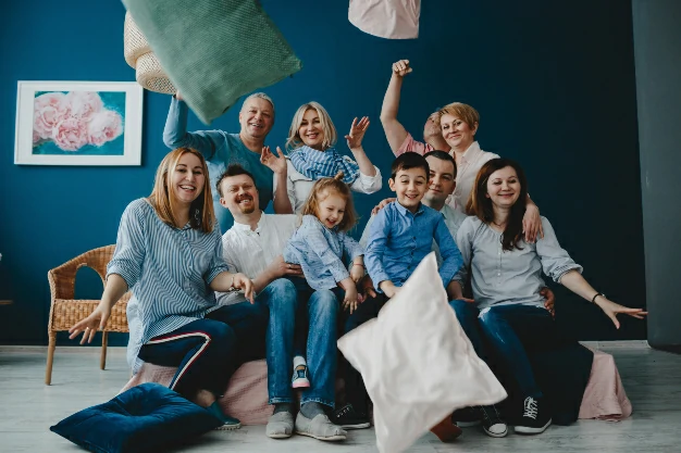 A imagem mostra uma família multigeracional composta por 10 pessoas, incluindo crianças, adultos e idosos, sentados e reunidos em torno de um sofá, jogando almofadas para o ar, simbolizando alegria e harmonia familiar. A imagem faz alusão a um dos serviços prestados na Clínica de Psicologia Elísios, ou seja, atendimento psicológico.